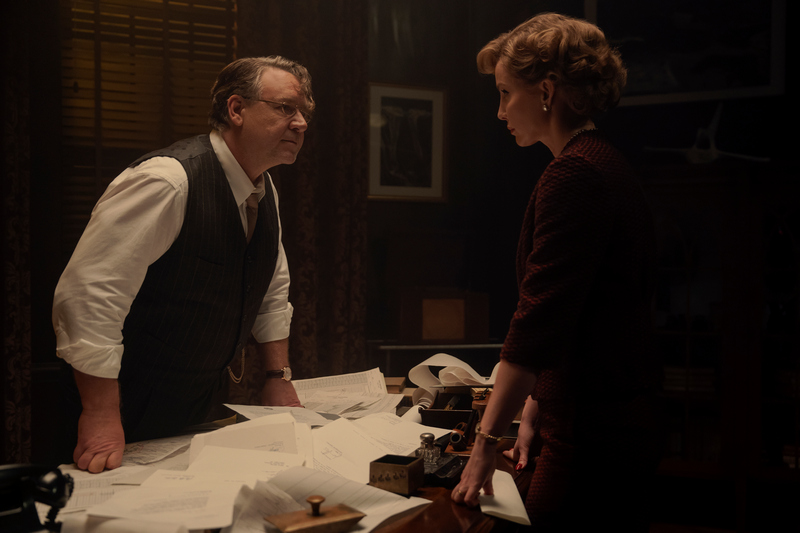 "A man and woman in formal attire have a serious discussion in a dimly lit office, standing over a desk covered with papers and old-fashioned equipment, creating a tense atmosphere."