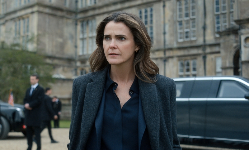 "A woman with wavy brown hair in a dark coat stands outside a grand historic building, looking serious. There is a black car and security personnel in the background, suggesting a formal or official environment."